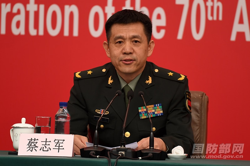 A Chinese man in uniform sitting in front of a red backdrop speaking into a mike.