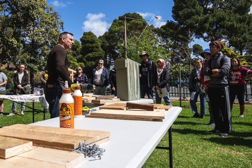 People stand about a wooden bird box on an outdoor bench.