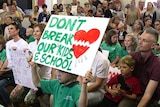A child holds a sign in crowded hall to fight a plan to radically reduce the size of the school.