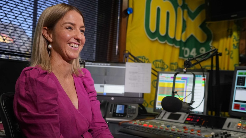 Katie Woolf is wearing a pink dress and smiling. She's in the radio studio.