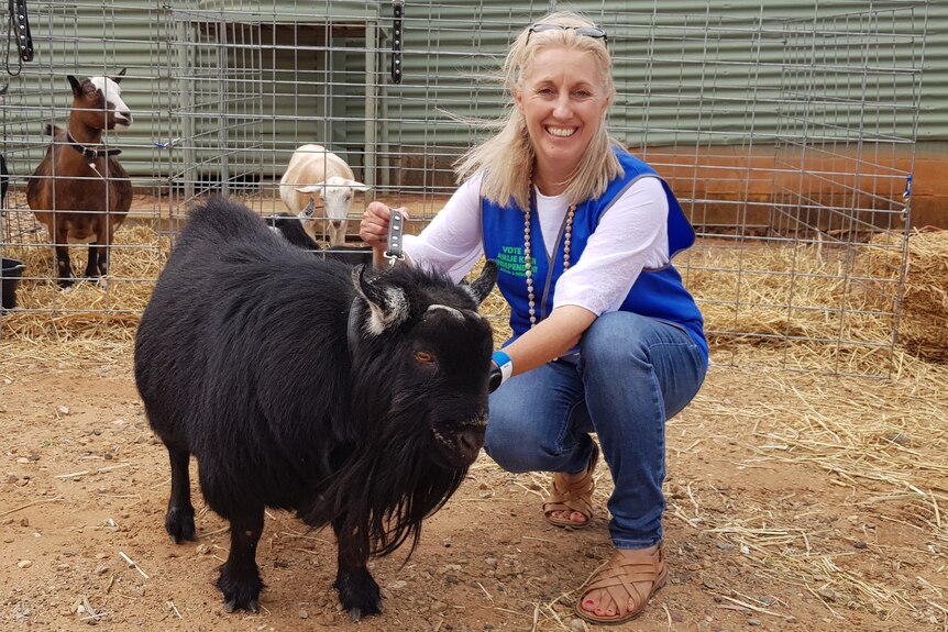 A blonde woman squatting next to a small black goat