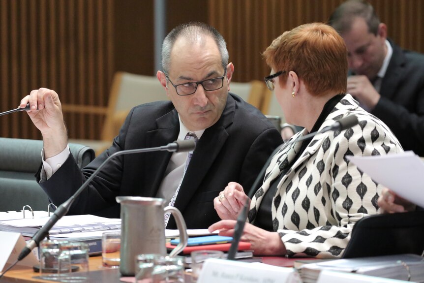 Mike Pezzullo covers his microphone while talking to Marise Payne at a table