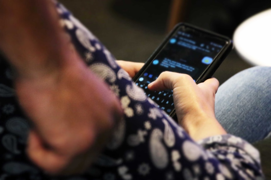 A women looks at her phone messages, as a man stands behind her with a clenched fist.