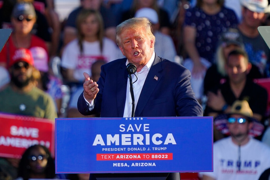 Donald Trump stands at a podium speaking with his hand up. Sign reads Save America. 