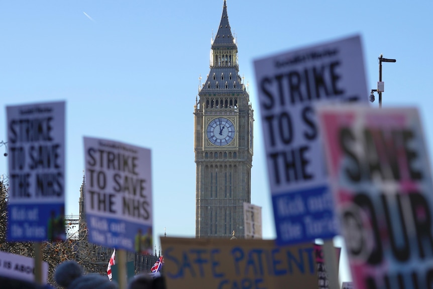 A massive clock on the side of a gold-trimmed tower is visible against a blue sky, with strike placards in the foreground.