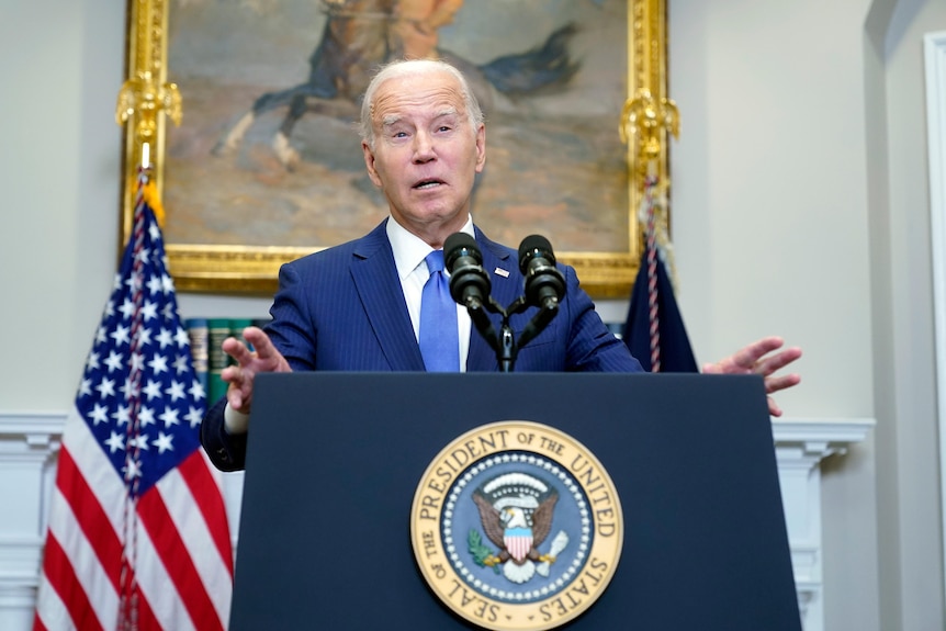 Joe Biden standing at a podium wearing a blue suit and the US flag behind him.
