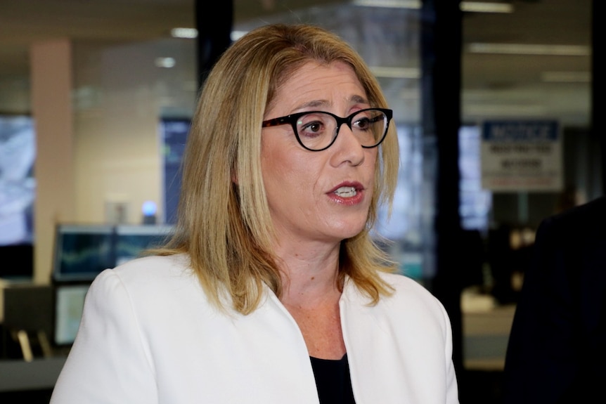 A woman with blonde hair wearing black-rimmed glasses and a white blazer.