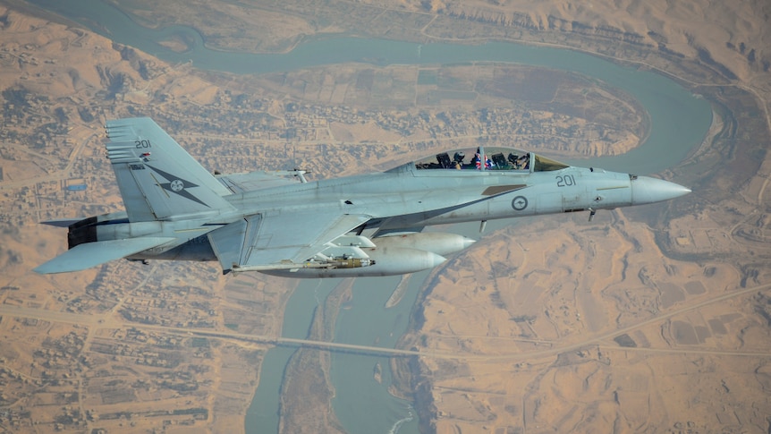 A military fighter aircraft with the RAAF kangaroo-in-a-circle logo flies above a river cutting through a dusty city.
