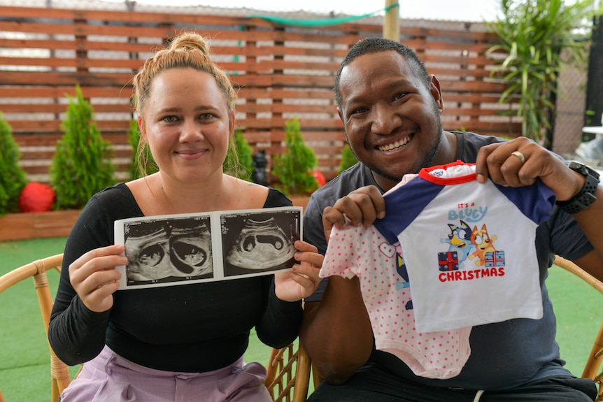 Tara and Luke Kaspar hold up two baby jumpsuits and a scan showing twins.