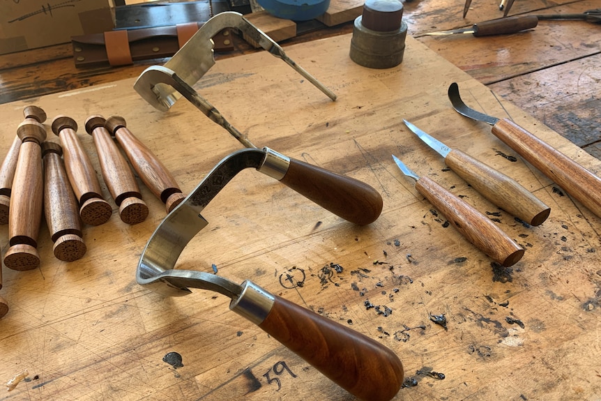 Six wooden handles, three finished knives and a scorp or double-handled blade tool on a bench.