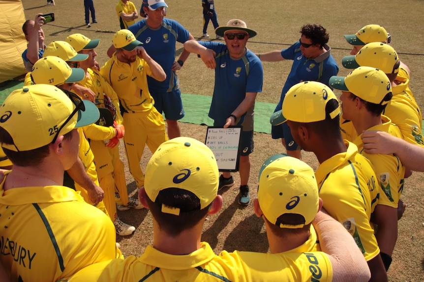 Jason Mathers talks to a group of players wearing yellow cricket kit