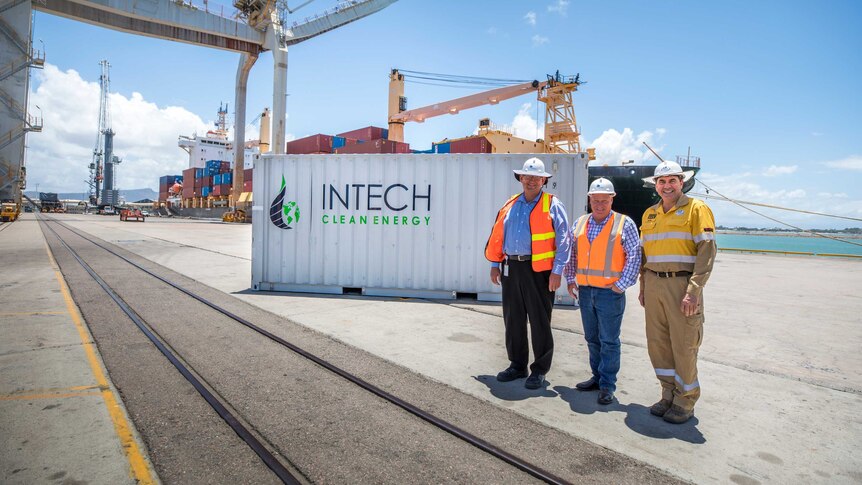 Three men wearing high vis and hard hats stand in front of a shipping container at a port