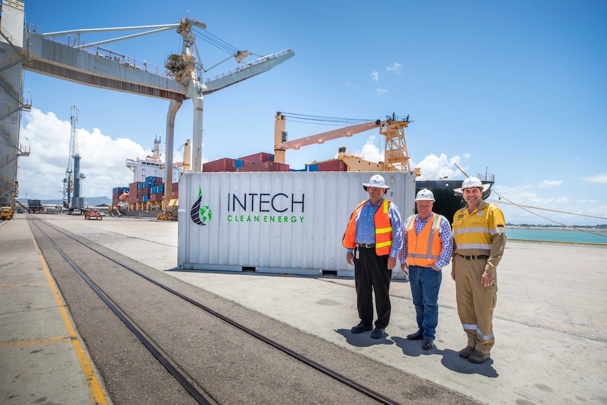 Three men wearing high vis and hard hats stand in front of a shipping container at a port