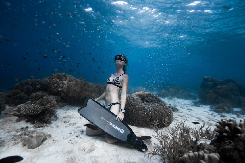 A woman in a bikini, flippers and snorkel mask sitting on the sand under shallow water.