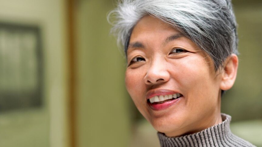 A woman with short grey hair and tan turtleneck jumper smiles widely.