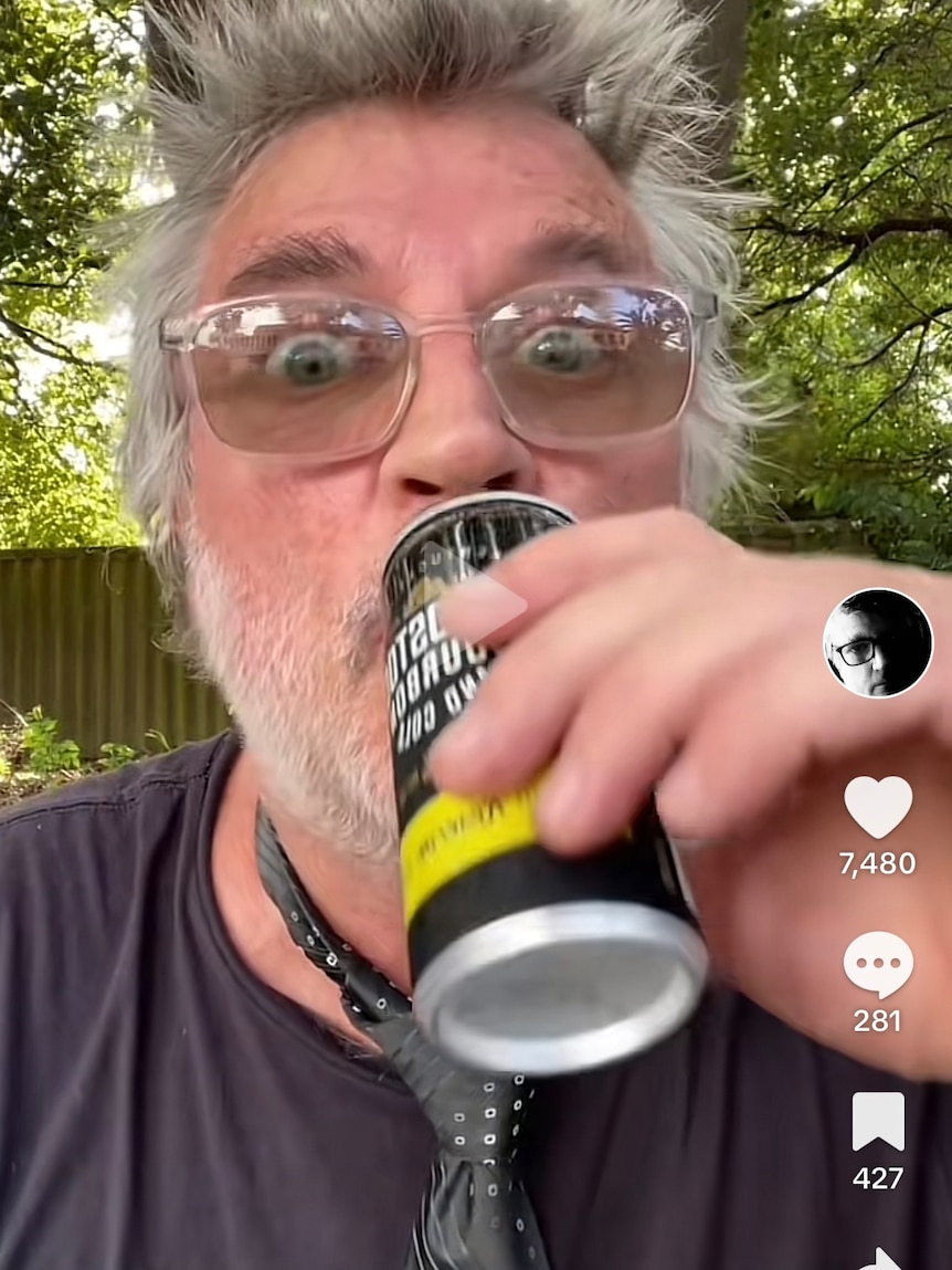 A man with spiked grey hair drinks from a can while taking a selfie.
