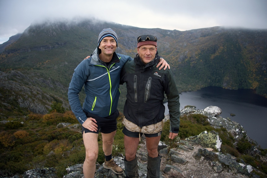 Two men wearing hiking gear, standing arm in arm with a misty vista of a mountain in the background. 