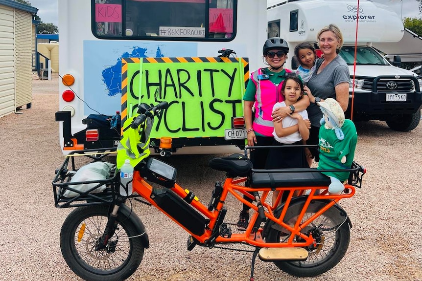 The two adults and two children stand near the bike and a motorhome with a 'charity cyclist' sign on the back