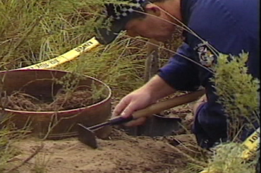 A forensic police officer uses a small tool to examine the ground in a bush setting.