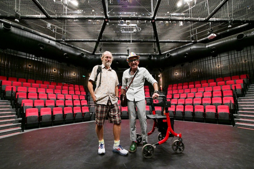 Two men stand on a black stage in front of red seating.