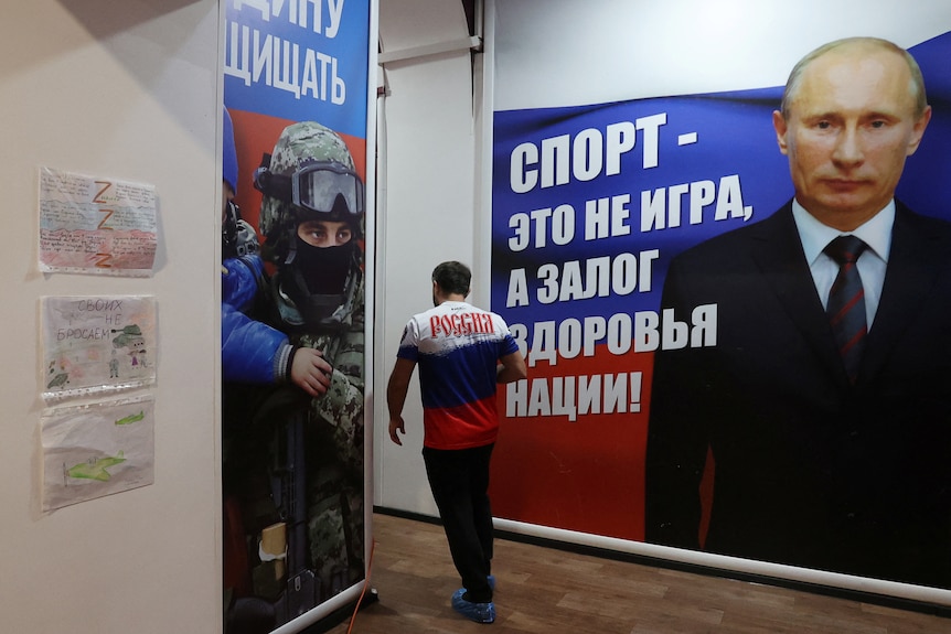 A man walks past a wall with a picture Mr Putin on it and large Russian writing. 
