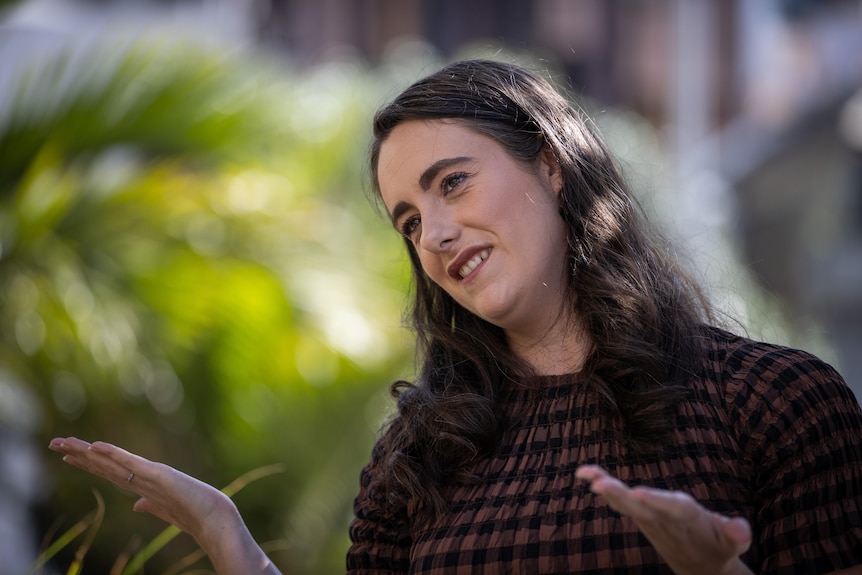 Dr Greaves wears a brown and black checked dress and her hair long as she gestures with her hands 