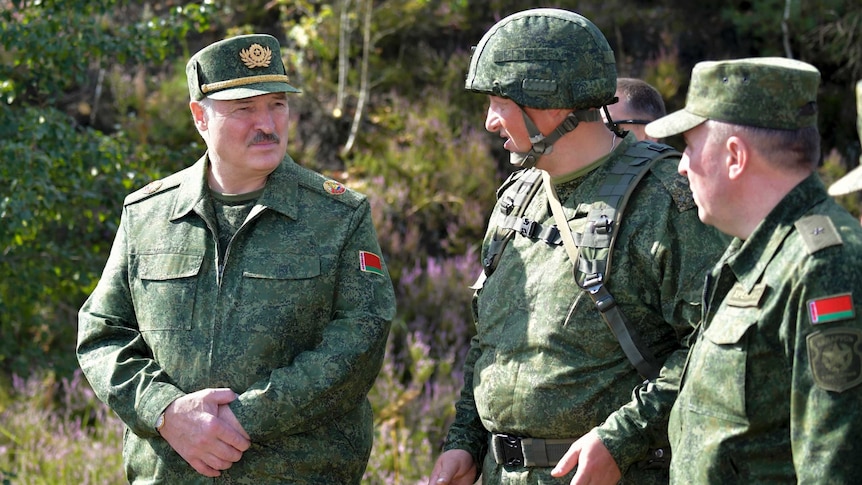 Three men in green military fatigues look at each other as they speak together while standing outside in sunshine.