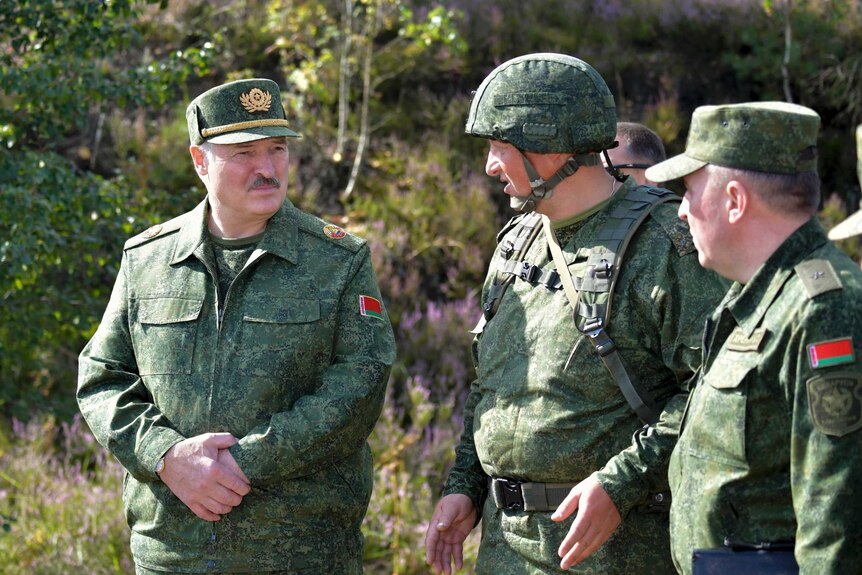 Three men in green military fatigues look at each other as they speak together while standing outside in sunshine.