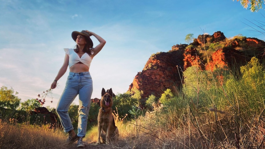 A young woman in a hat, white top and jeans stands next to a dog in an outback setting
