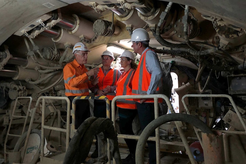 Four people in high viz and hard hats stand on the boring machine that is building a tunnel.