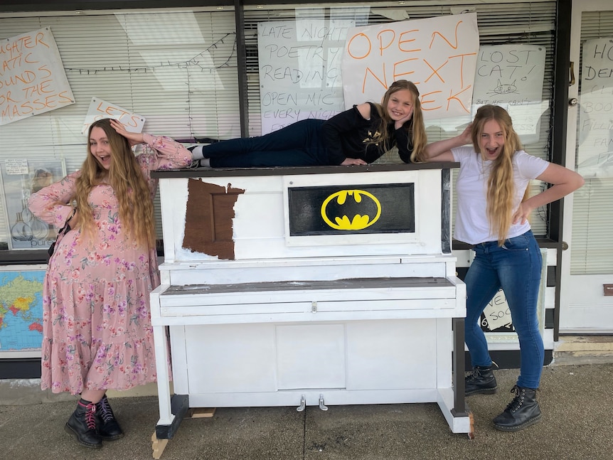 Three girls posing in an exaggerated way around a white piano sitting on a pavement outside a shopfront.