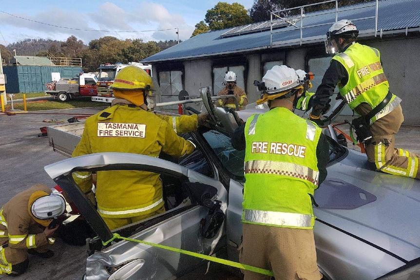 Six firefighters in high-vis and helmets working to cut open a silver car in crash rescue training.