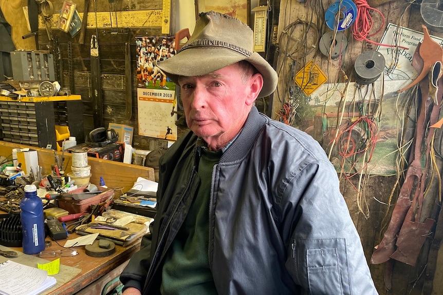 An older man wearing a worn Akubra hat sits in a workshop, surrounded by tools and materials.