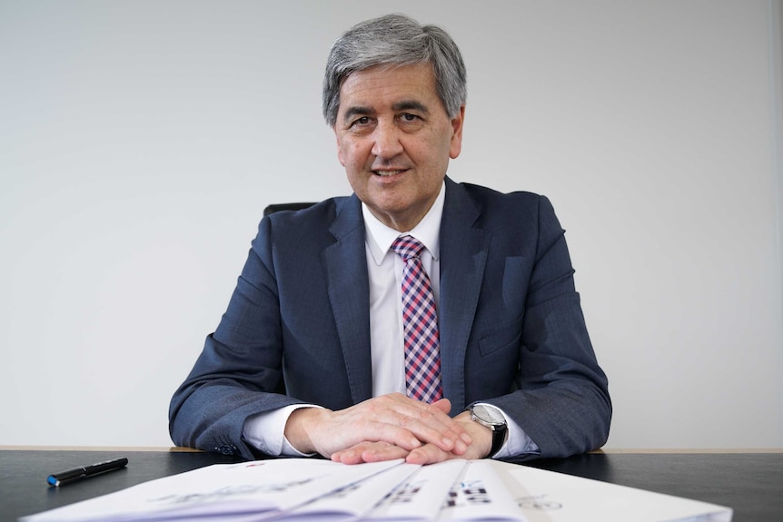 A man with grey hair, in a suit and tie, seated at a table with budget papers and pens.