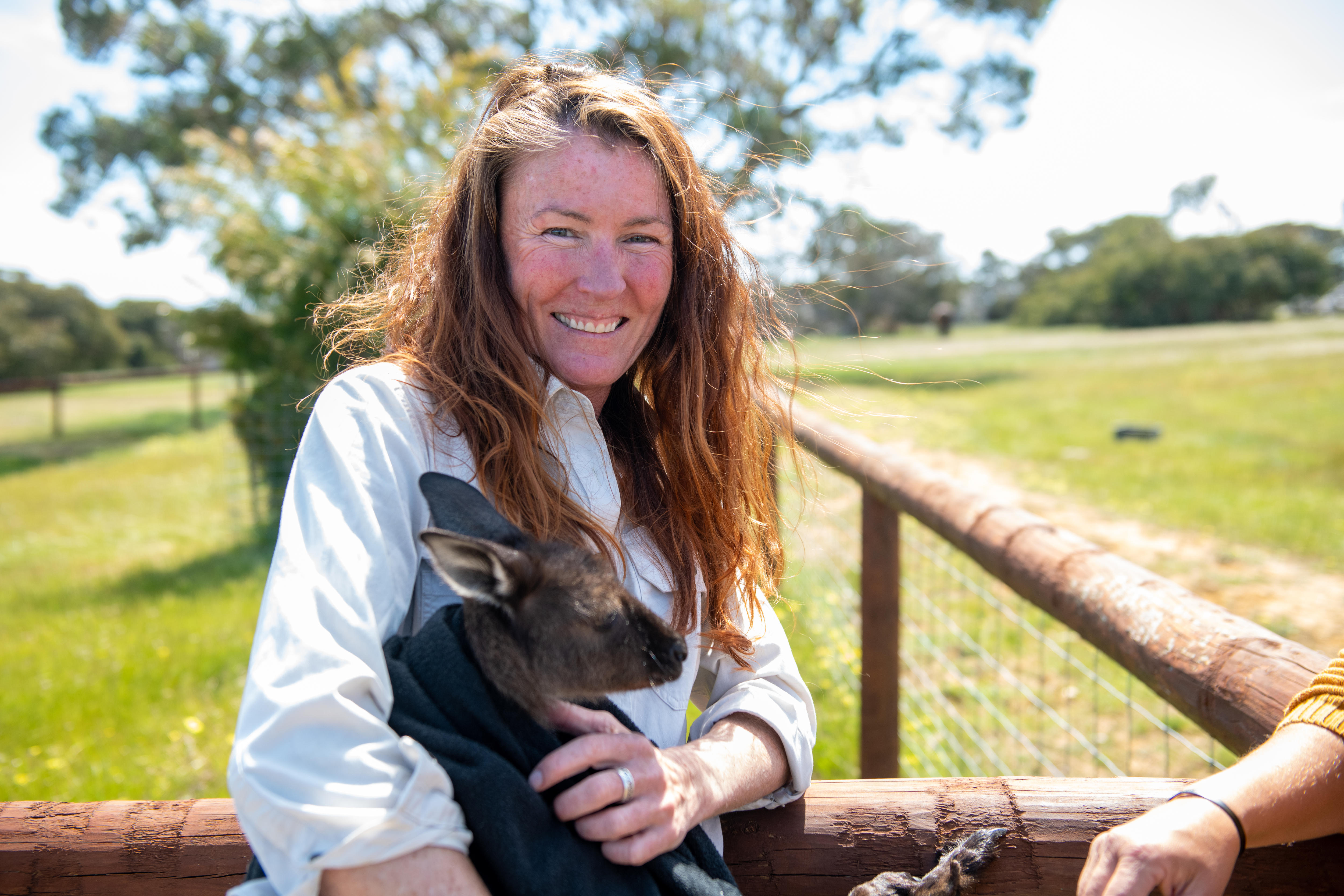 Woman smiling holding a kangaroo in a blanket, standing in a meadow. 