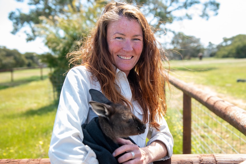 Woman smiling holding a kangaroo in a blanket, standing in a meadow. 