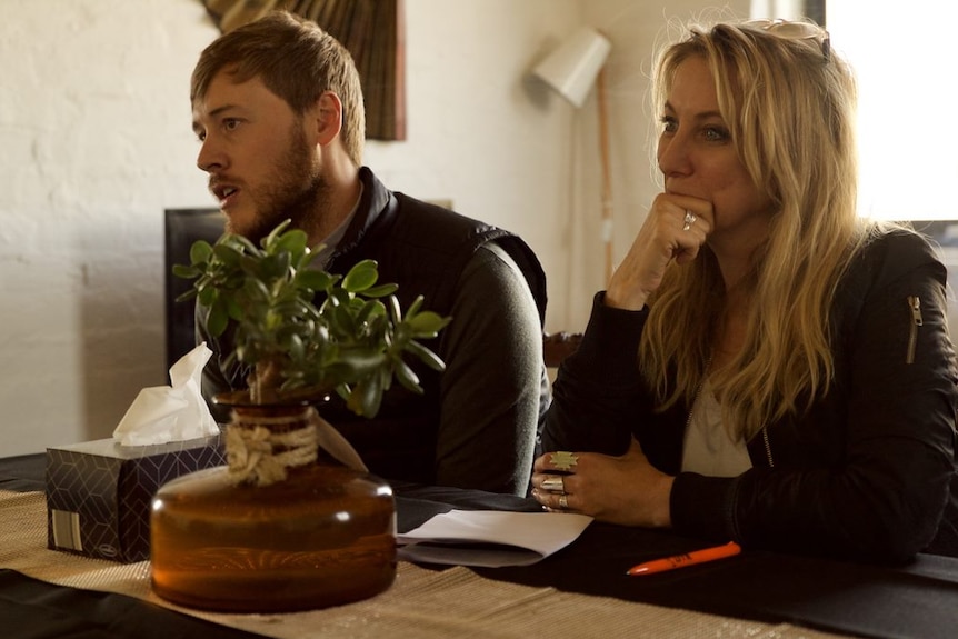 A young man and a woman with long blonde hair sit beside each other at a coffee table. They are in conversation with someone.