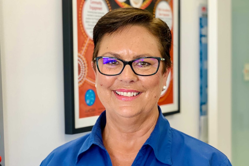 A head shot of a smiling short-haired woman in blue shirt, black rim glasses standing in front of an Indigenous print.