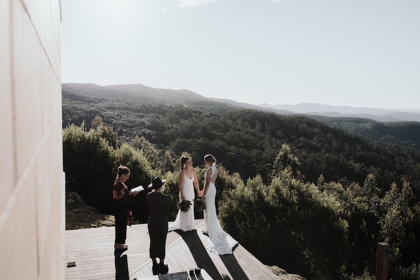 Two women in bridal gowns say vows with a celebrant in front of a spectacular bush backdrop.