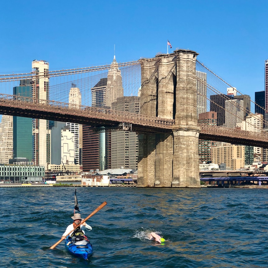 NYC's infamous Brooklyn Bridge on a sunny day while underneath there is a kayak and a man swimming