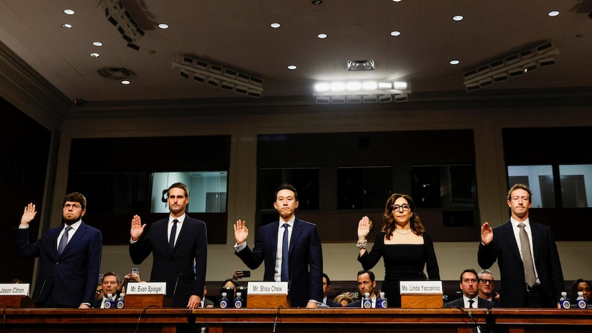 Five people wearing black suits and clothes lifting their right arm up to swear while standing behind a brown desk 