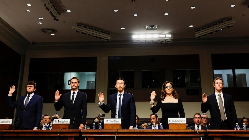 Five people wearing black suits and clothes lifting their right arm up to swear while standing behind a brown desk