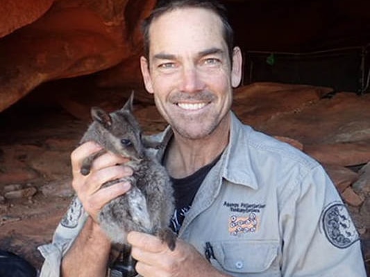 A man with brown hair holds a wallaby and stands near a rock shelter