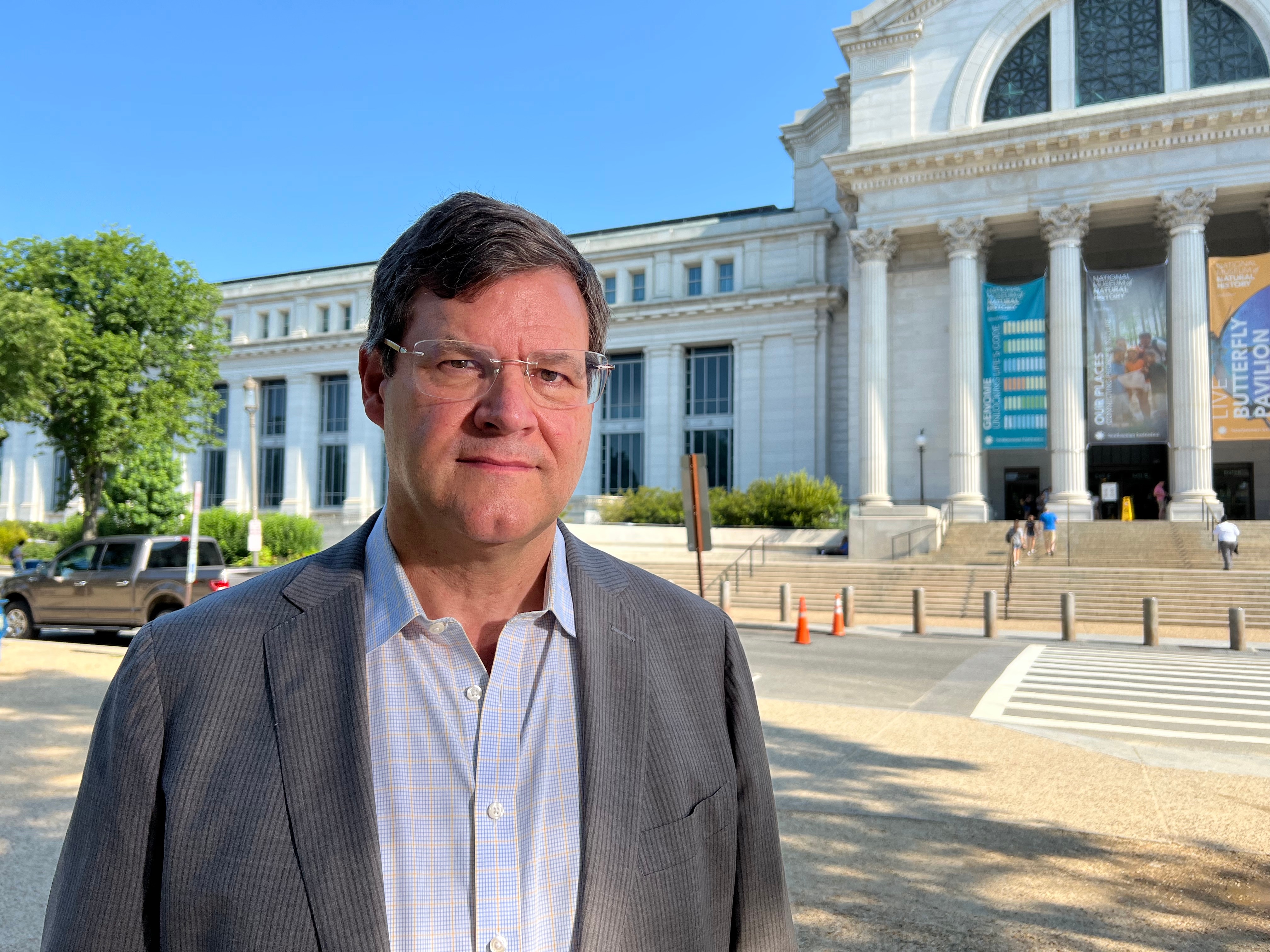 A man with glasses in a white shirt and gray jacket stands in front of the Smithsonian