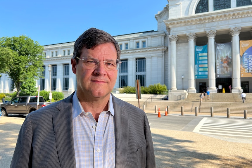 A man with glasses in a white shirt and grey jacket stands in front of the Smithsonian