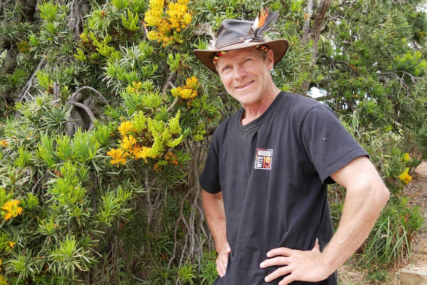 A man wearing a hat stands next to a tree with yellow flowers.