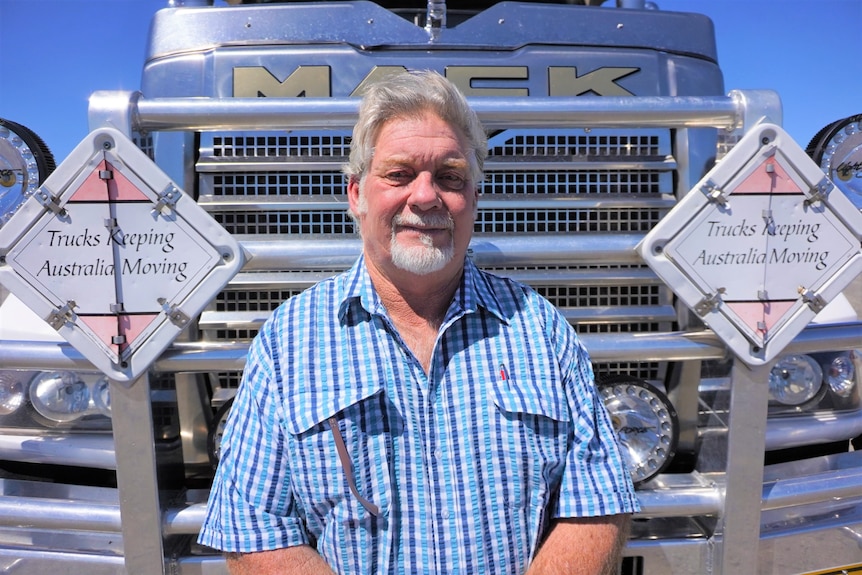 A man wearing a blue checked shirt stands in front of a B-double truck