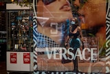 A woman wearing a mask and walking, seen through the reflection of a window featuring an ad for Versace sunglasses.