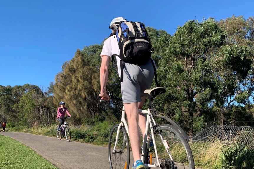 A man on a bicycle pedals past a women on a bicycle on a paved pathway that runs through a park.