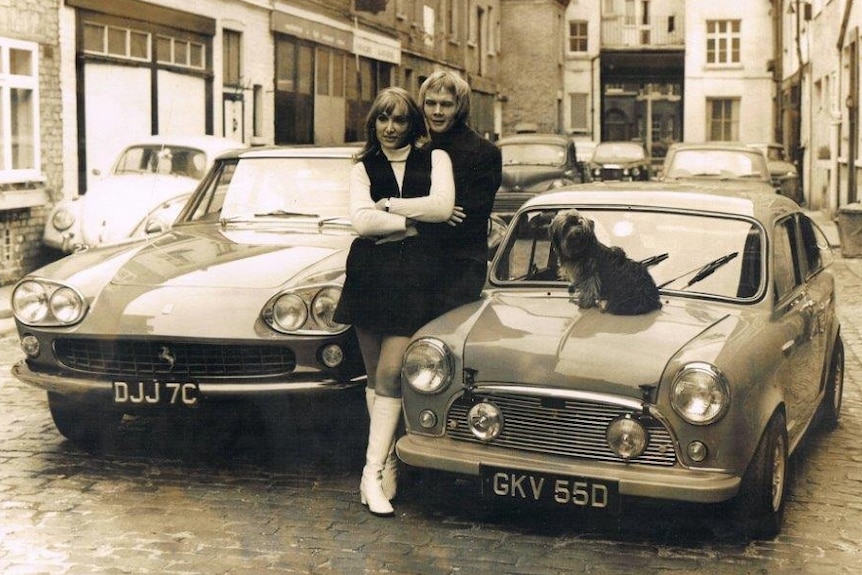 A man and woman stand next to two cars in a London Street in the sixties